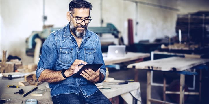 Mann sitzt auf Werkbank und schaut ins iPad Mann sitzt auf Werkbank und schaut ins iPad